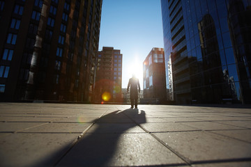 Silhouette of businessman wearing coat standing at glass office buildings and looking at camera, lens flare