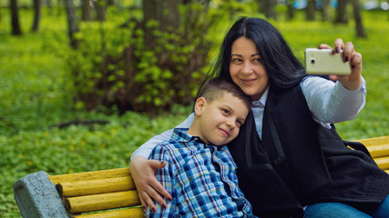 Naklejka premium Mom and son in the park on a yellow bench are photographed selfies on a mobile phone. Family photo session.