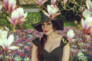 Fototapeta na wymiar Beautiful girl with black hair in a black dress on a background of a magnolia flower. Blurring background. Shooting outdoors. The concept of fashion and beauty. Beautiful girl and magnolia flowers. 