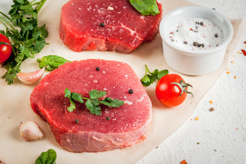 Raw beef, steak, cutlet. Meat, a source of protein. On a cutting board, on a white stone table. With spices, herbs and tomatoes for cooking. Copy space