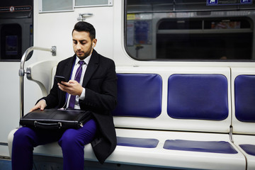 Portrait of Middle-Eastern businessman commuting to work in subway train, using smartphone to...