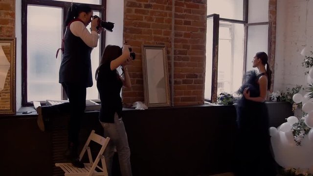 Many photographers on the masterclass take pictures of a beautiful model in a black wedding dress on the background of a red brick wall by the window. Backstage photoshoot.