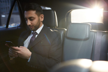 Portrait of handsome Middle-Eastern businessman using smartphone on backseat inside expensive car
