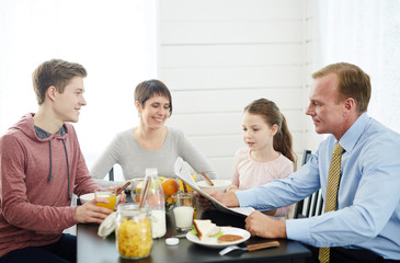 Family of four sitting around kitchen table and enjoying delicious breakfast, concentrated father reading newspaper together with his curious little daughter