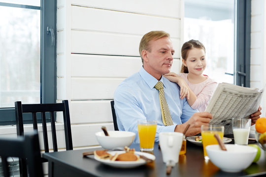 Fair-haired middle-aged man and his pretty little daughter wrapped up in reading newspaper article while having tasty breakfast in dining room, waist-up portrait