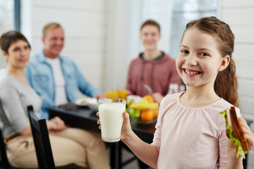 Cute little girl with ponytail holding glass of milk and tasty sandwich in hands while looking at camera with toothy smile, her family sitting at kitchen table