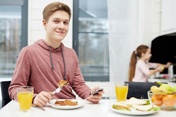 Obraz na płótnie Canvas Waist-up portrait of handsome teenage boy looking at camera with wide smile while eating tasty pancakes at breakfast, his little sister standing at sink and washing dishes