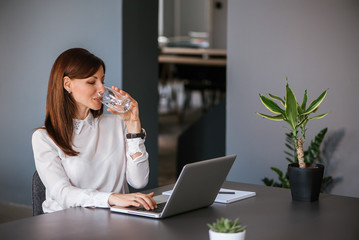 Stay hydrated. Pretty young woman in the office drinking water while working. Drinking from glass.