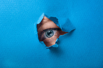 A female eye looks through a hole in a paper blue background.
