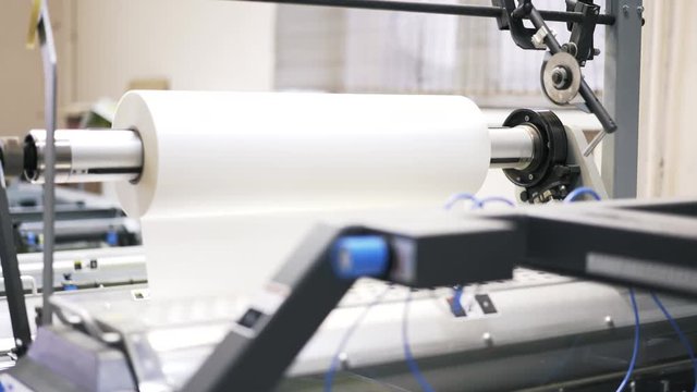Large roll of white paper is rotating at a printing plant machine. Locked down real time medium shot