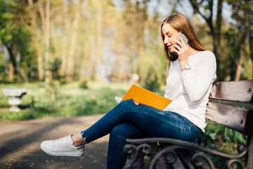Portrait of attractive young professional woman using a smartphone while sitting on a wooden bench in a park, smiling.