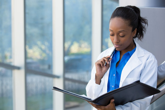 Closeup portrait of friendly, thinking confident female doctor, healthcare professional with labcoat, holding pen to face and holding notebook pad. Isolated hospital clinic background.