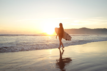 Young female surfer entering the ocean with surfboard at sunset or sunrise