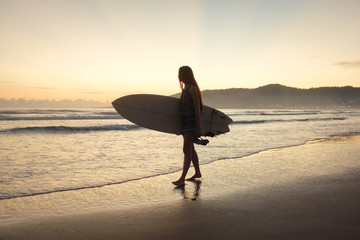 Fototapeta na wymiar Female surfer with surfboard looking into the ocean during sunset sunrise