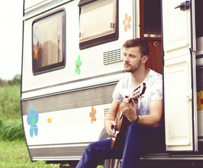 Young, handsome, bearded hipster playing guitar outdoors at summer. Holiday, journey, vacation concept.