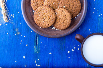 Oatmeal cookies in brown plate near cup of milk on blue background. Rustic style. Copy space. Top view