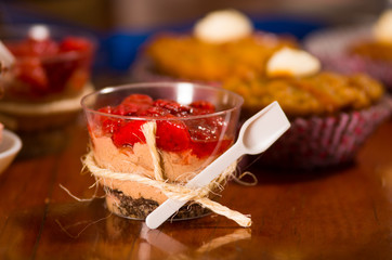Delicious mousse covered in strawberry topping, presented inside small glasses with spoons attached, cake pieces and muffins background, pastry concept