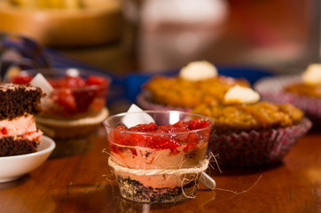 Delicious mousse covered in strawberry topping, presented inside small glasses with spoons attached, cake pieces and muffins background, pastry concept