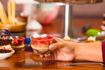 Hand grabbing onto delicious mousse covered in strawberry topping, presented inside small glass, cake pieces and muffins background, pastry concept