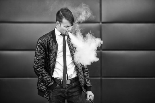 Vape man. Portrait of a handsome young white guy with modern haircut vaping and letting off steam from an electronic cigarette. Black and white photo. Lifestyle.