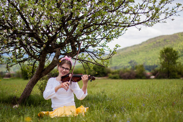 Beautiful blonde girl sitting on a beautiful green grass full of flowers and enjoy the sounds of the violin, girl playing the violin in the countryside on a beautiful meadow