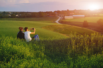 young loving couple sits on a hill with phone on the green field background. couple makes selfie. romantic date at evening