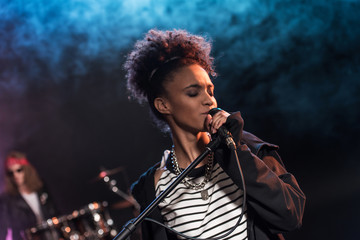 Female singer with microphone and rock and roll band performing hard rock music on stage - obrazy, fototapety, plakaty