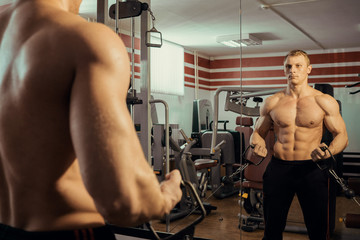 Naklejka premium Young strong athlete with a beautiful naked torso doing muscle exercises in the gym in the mirror
