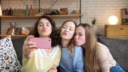 three young female best friend have fun and make selfie, grimacing and laughing using telephone mobile camera, while sitting at home on sofa