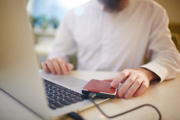Close-up shot of unrecognizable businessman in buttoned-up white shirt sitting at cafe table and using laptop connected to external hard drive