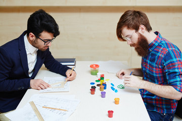 Profile view of concentrated young architect in suit making changes to blueprint while other man...