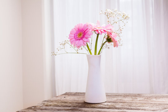 vacker bukett med rosa gerbera i vit vas på trä bord och vit gardin i bakgrunden