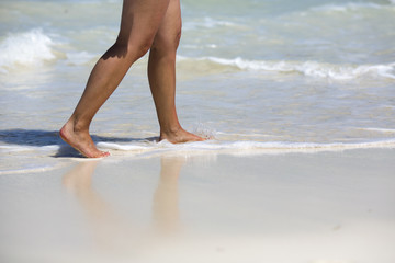 Caucasian tanned woman is walking on the beach. Water and sand on the feet. White sand beach with waves.