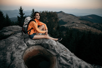 Naklejka na ściany i meble Young woman backpacker hiking on mountain peak cliff. Young woman sitting on a rock with backpack and looking to the horizon. Hipster young girl with backpack enjoying sunset on peak mountain