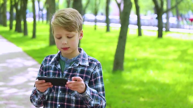 Portrait of blond cute boy of 10 year old standing in spring or summer urban park and listening to music happily. Cheerful kid uses black headphones and modern smartphone. Real time video footage.