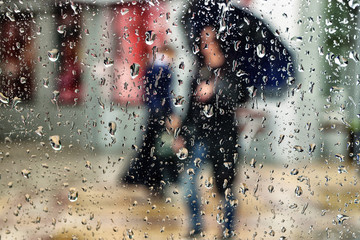 View through sharp raindrops on the window glass at the girl in a black jacket with black umbrella in her hand and going outside in the rain