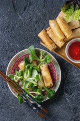 Fried spring rolls with red pepper sauces, served in traditional china plate with fresh green salad and wooden chopsticks over black texture background. Flat lay, space. Asian food