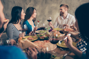 Young handsome bearded guy is telling the toast to a brunette cute girl, a birthday girl. Friends are celebrating with tasty dishes and drinks, smiling and enjoying themselves
