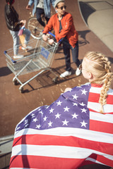 Blonde young girl with american flag looking at friends having fun at skateboard park