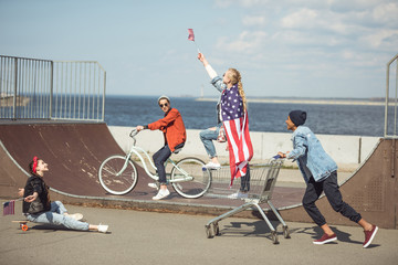 Obraz premium Teenagers group with skateboard, bicycle and shopping cart having fun together and waving american flags at skateboard park