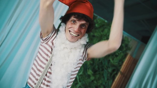 Joyful caucasian man in dwarf costume white beard, red cap striped shirt suspenders smiling lifting old weary broken tin tuba above head