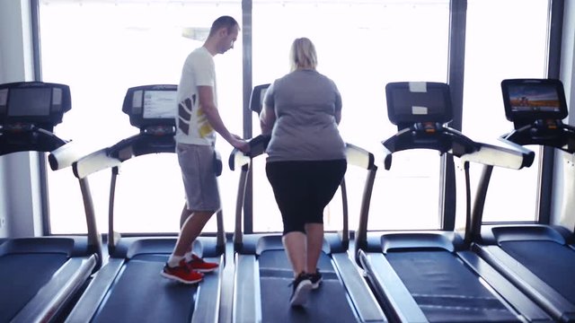 Overweight woman with trainer walking on treadmill in gym.