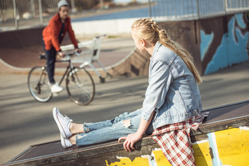 hipster girl resting at skateboard park while boy riding bicycle, teenagers having fun concept