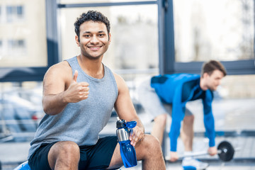 Handsome young man with bottle of water resting at gym, other man workout on blurred background