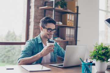 Obraz premium Smiling handsome worker in glasses drinking coffee while watching video on his laptop at office