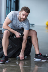 Young man sitting on tyre and exercising with small dumbbells