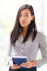 Waist-up portrait of confident Asian businesswoman looking at camera while standing against blurred background with notebook in hands