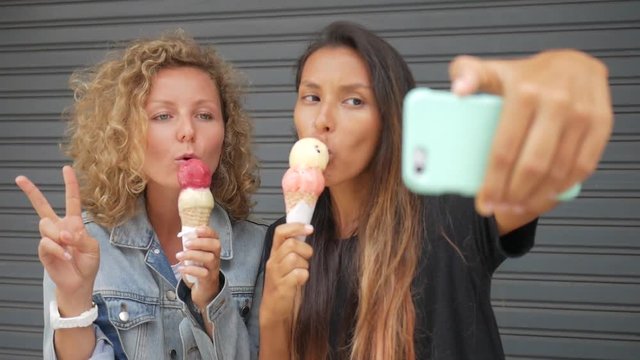Two Young Carefree Mixed Race Hipster Girls Eating Ice Cream and Posing for a Selfie Photo using Mobile Phone. Lifestyle HD Slowmotion Footage.