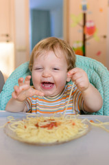 Cute one year old baby eats spaghetti in a children's chair.