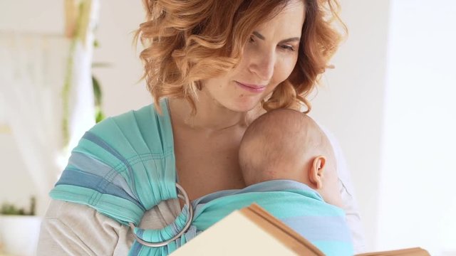 Beautiful middle-aged Caucasian female reading book while her newborn son sleeping in baby sling. Close up footage in slowmotion